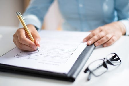 Person in blue shirt writing on a form with a gold pen at a desk with glasses nearby